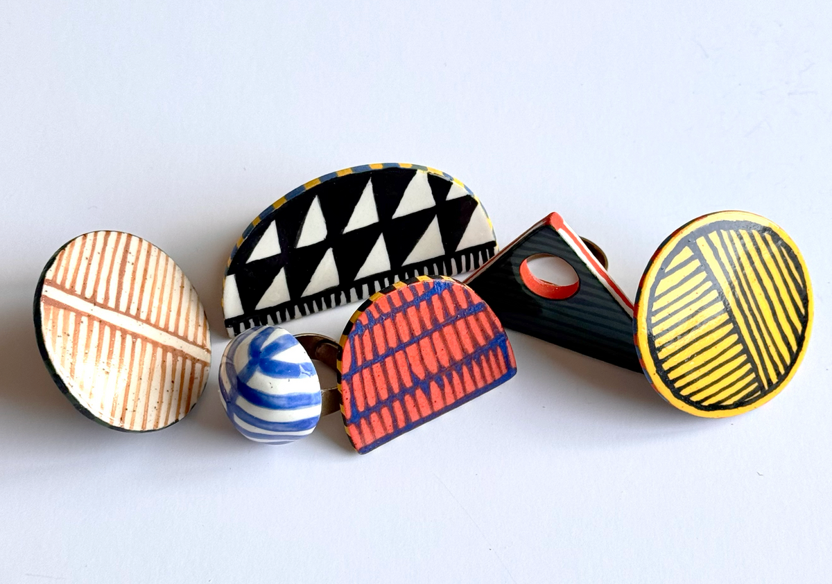 A set of five colorful, handcrafted wooden geometric pins arranged on a plain white background. The designs include striped, triangular, wavy, and textured wood patterns in black, yellow, blue, red, and beige.